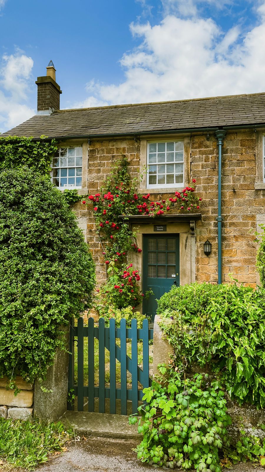 A honey-stone English cottage with climbing roses and a teal picket gate
